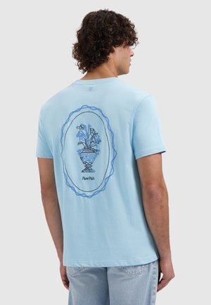 Man met krullend haar die een lichtblauw T-shirt draagt met een geborduurde blauwe bloemenvaas en de tekst "Pure Path" op de achterkant.