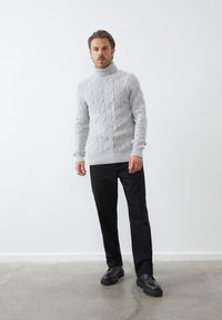 Jasnoszary sweter typu golf z dzianiny w splot warkoczowy, z prążkowanymi mankietami i dołem, zestawiony z czarnymi spodniami i czarnymi butami, na jednolitym tle.