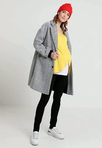 Mulher em pé a usar um gorro vermelho, um casaco cinza com padrão, uma blusa amarela, calças pretas e ténis brancos, contra um fundo claro e liso.