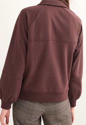 Vrouw gekleed in een bruin longsleeve shirt met pofmouwen en een grijze geruite broek, gezien van achteren.