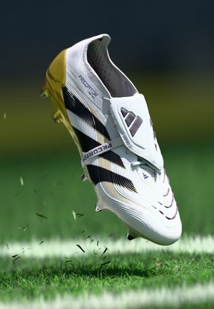 Scarpa da calcio bianca con strisce nere e dettagli dorati, dotata di una parte superiore testurizzata, design flessibile e un'ampia fascetta sopra il collo del piede.