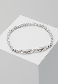 Srebrna bransoletka z rzędem okrągłych przezroczystych kamieni osadzonych w metalu. Widoczny detal zapięcia, z połyskliwym wykończeniem i gładką teksturą.