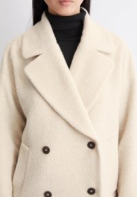 Dobbeltbrystet cremede frakke lavet af struktureret stof med store sorte knapper og en notched krave, lagdelt over en sort turtleneck.