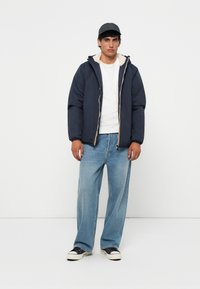 Navy puffer jacket with a cream fleece lining, paired with light blue baggy jeans and black sneakers. Cap worn backward, minimalist design.