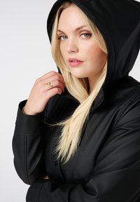 Zwarte hoodie met een gladde afwerking en ritssluiting. Model houdt hand bij de nek, met een gouden ring om. Licht haar contrasteert met de donkere stof.