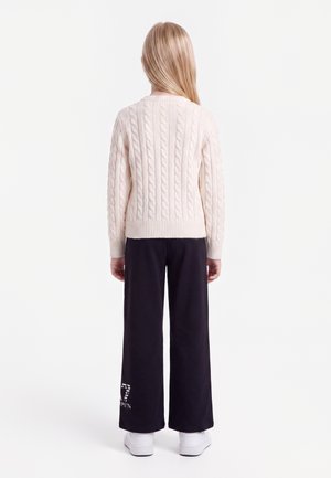 Kind mit langen blonden Haaren, das einen cremefarbenen Zopfpullover, schwarze weite Hosen mit weißem Text und weiße Turnschuhe trägt, von hinten gesehen.