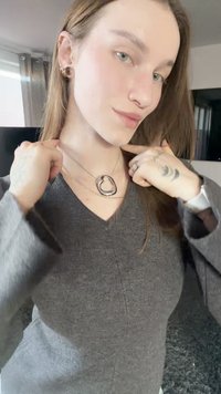 Grijs v-hals trui, zilveren gelaagde ketting met een hanger in organische vorm, korte mouwen, en een close-up van tatoeages op de handen.