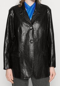 Blazer en similicuir noir à texture crocodile, doté d'un col cranté, de deux boutons et de poches latérales, associé à un haut bleu.