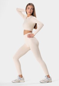 Béžový dlhý rukáv crop top a legíny s vysokým pásom a jemnou textúrou. Modelka v svetlých teniskách, pózujúca na jednofarebnom pozadí.
