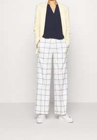 Beige blazer over een marineblauwe V-hals top, gecombineerd met witte geruite broek met blauwe lijnen en witte sneakers met tekstuurzolen.