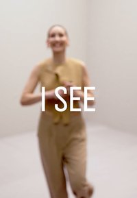 Beige jumpsuit med ermeløs topp og knytedetalj ved halsen. Bakgrunnen er en lys, nøytral farge. Tekst over: "JEG SER."