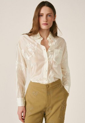 Mujer con una camisa blanca transparente bordada, abotonada y metida dentro de pantalones de tiro alto color beige con bordados florales, de pie con una mano en el bolsillo.