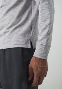 Grå långärmad skjorta i strukturerad tyg, med sidoflikar vid nederkanten. Skjortan är kombinerad med mörka shorts.