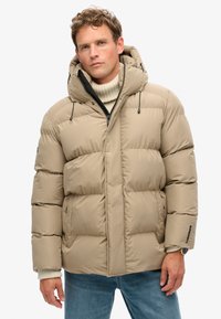 Giacca imbottita beige con cappuccio, dotata di chiusura con zip, coulisse e tessuto testurizzato. Le sezioni cucite creano un aspetto gonfio.