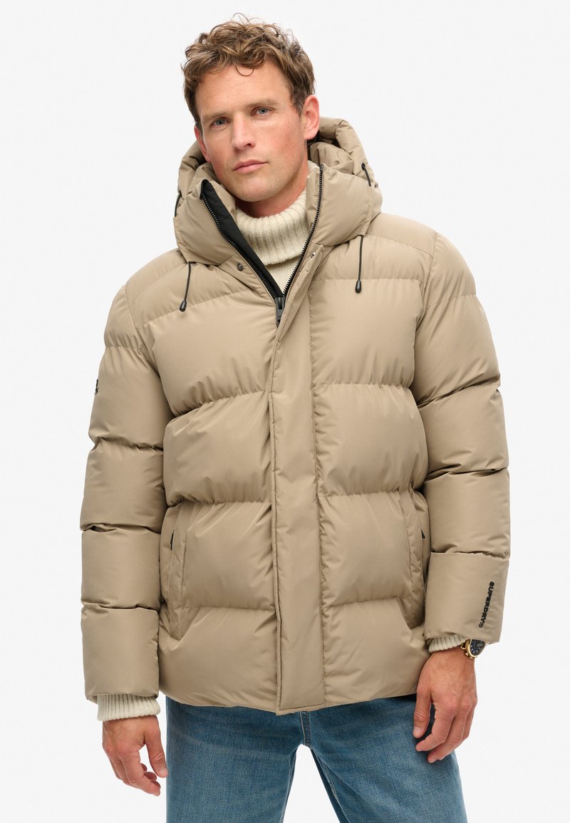 Giacca imbottita beige con cappuccio, dotata di chiusura con zip, coulisse e tessuto testurizzato. Le sezioni cucite creano un aspetto gonfio.