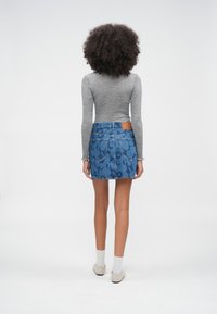 Göndör hajú személy háttal áll, szürke hosszú ujjú felsőt, kék virágmintás farmerszoknyát, fehér zoknit és fehér cipőt visel.