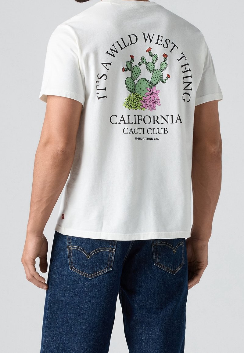 T-shirt blanc avec motif cactus et fleurs, texte « It’s a Wild West Thing, California Cacti Club, Joshua Tree CA », porté par une personne en jean bleu.