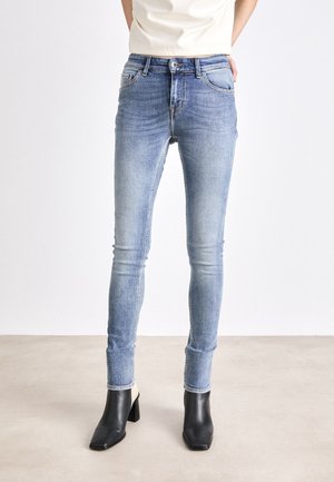 Personne portant un jean skinny bleu clair et des bottines noires à talons, debout sur un sol clair contre un mur blanc uni.