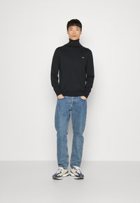 Svart polotröja, ljusblå denimjeans, vita och marinblå sneakers med en logotyp på vänster bröst; slät textur och figursydd design.
