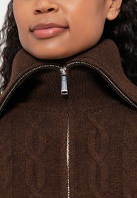 Pruun kootud kampsun kõrge kaelusega ja hõbedase tõmblukuga. Iseloomustab tekstureeritud kabelmustrit, mis näitab pehmet ja sooja materjali.