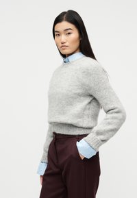 Γυναίκα με μακριά σκούρα μαλλιά, που φοράει ανοιχτό γκρι χνουδωτό πουλόβερ πάνω από μπλε πουκάμισο με κουμπιά και σκούρο μπορντό παντελόνι, στέκεται με το ένα χέρι στην τσέπη.