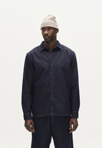 Camisa de mezclilla oscura con cuello extendido, que cuenta con mangas largas y botones negros. Combinada con pantalones oscuros a juego y un gorro de lana beige.