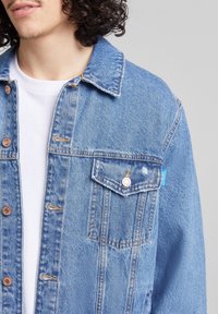 Light blue denim jacket with a relaxed fit, metal snap buttons, two chest pockets, and orange stitching details. White t-shirt underneath.