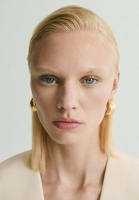 Modelo con piel clara y cabello rubio claro y lacio, que lleva grandes pendientes circulares dorados, un maquillaje mínimo y una prenda beige.
