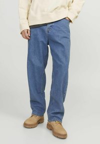 Moški nosi ohlapne modre kavbojke, bež pulover in svetlobrne čevlje z vezalkami, stoji ob enotnem svetlem ozadju.