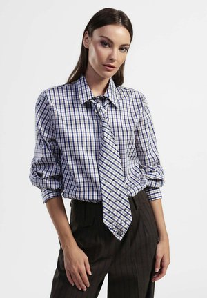 Camicia a quadri blu e bianca con cravatta annodata, realizzata in tessuto liscio e con le maniche arrotolate. Abbinata a pantaloni scuri a righe sottili.