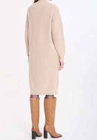 Robe beige en maille à col montant et manches longues, tombant jusqu'au milieu du mollet, associée à des bottes en cuir marron montantes jusqu'au genou et des collants noirs.