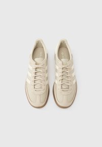 Béžové tenisky s třemi bílými pruhy, svrškem z texturované kůže a hnědou gumovou podrážkou. Mají tradiční šněrování a detaily s logem.