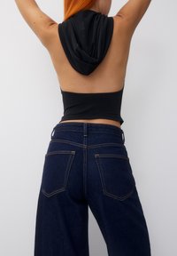 Top sans manches noir avec capuche et dos ouvert, associé à un jean ample bleu foncé avec poches arrière et coutures contrastées.