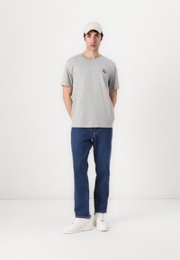 Tricou gri cu mânecă scurtă, având un mic logo albastru, asortat cu blugi albaștri și adidași albi, completat cu o șapcă bej.