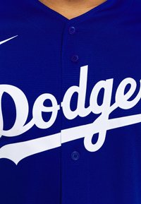 Modrá baseballová dres s bílým nápisem "Dodgers" přes přední stranu, se dvěma knoflíky a strukturou látky. Logo Nike na rameni.