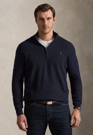 Mand iført en marineblå sweater med kvart-lynlås over en hvid skjorte, mørke jeans med et brunt bælte og et armbåndsur, stående mod en neutral baggrund.