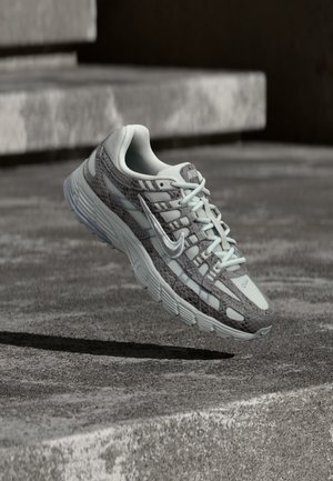 Chaussure de course Nike grise et blanche avec motif texturé, laçage avant et semelle épaisse, flottant au-dessus d'une marche en béton en extérieur.