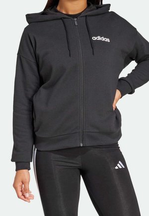Person iført en sort Adidas lynlåshoodie med hætte og sorte Adidas leggings med hvide striber og logo på venstre lår.