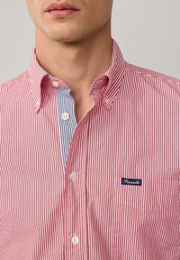 Rot-weißer gestreifter Hemd mit Knopfleiste und Schalkragen, ausgestattet mit einer blau-weiß gestreiften inneren Leiste und einem kleinen Logo-Patch auf der Brust.