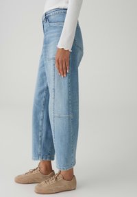 Persona che indossa jeans patchwork azzurri, maglia bianca a maniche lunghe e sneakers in suede beige, in piedi di lato su uno sfondo semplice.