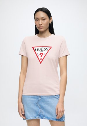 Kvinde med langt sort hår iført en lyserød Guess t-shirt med en rød trekantslogo og en lyseblå denim nederdel, der står mod en ensfarvet baggrund.