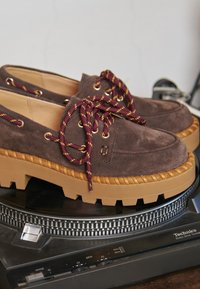 Scarpe in suede marrone con suola in gomma beige spessa e battistrada profondo. Presentano lacci intrecciati color bordeaux e oro, occhielli dorati e un design mocassino.