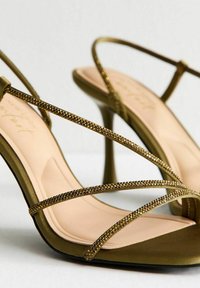 Sandali con tacco alto verde oliva, caratterizzati da fasce incrociate decorate con piccoli accenti metallici. Rivestimento interno liscio e design della suola sagomato.