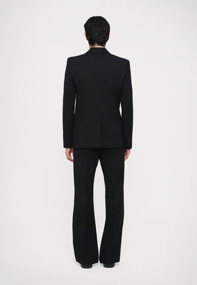 Costum negru la comandă, compus dintr-un sacou ajustat cu rever în formă de clapă și pantaloni evazați. Țesătură netedă cu un finisaj mat, fără modele sau accente vizibile.