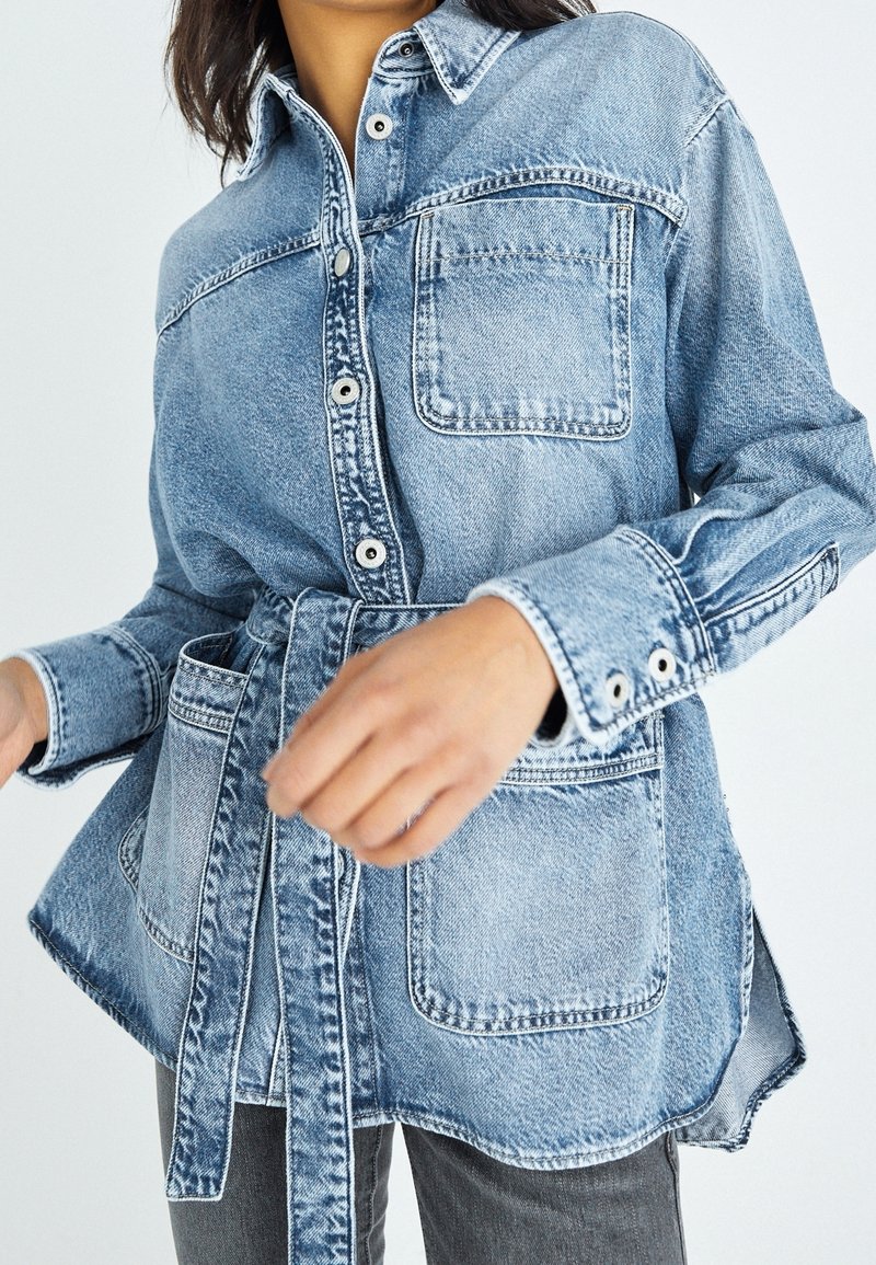 Denimskjorta med ljusblå tvätt, knäppning fram, krage och långa ärmar. Utrustad med midjebälte och två bröstfickor.