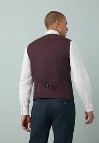 Gilet bordeaux foncé au design élégant, avec un détail de boucle à l'arrière et une texture de tissu lisse, porté sur une chemise blanche.