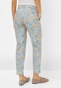 Geprinte broeken met een beigen basis en blauwe bloemenpatronen. Slim fit met zakken en een rechte pijp. Platte schoenen zichtbaar.