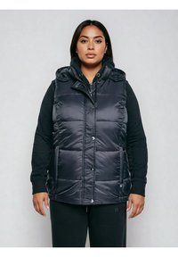 Naine kannab musta kapuutsiga pufferi liivakut musta pikkade varrukatega kapuutsiga jope peal, seistes ühevärvilise helehalli tausta ees.