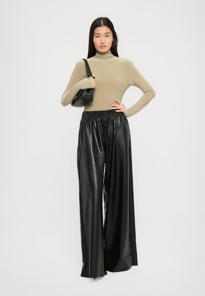 Donna che indossa un dolcevita beige e pantaloni in pelle neri a gamba larga, con una borsa nera appesa alla spalla, davanti a uno sfondo neutro.