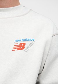 Ανοιχτογκρι φούτερ με μπλε κείμενο "new balance", κόκκινο λογότυπο "NB" και γκρι κείμενο "RUNNING" στο στήθος.